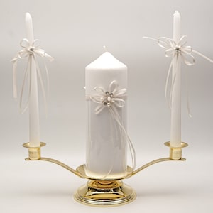 May include: A gold metal candle holder with three white candles. The center candle is larger than the two side candles. Each candle has a white satin ribbon tied around it with a small pearl and rhinestone embellishment.