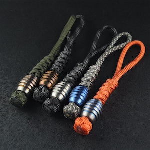 May include: Five beaded lanyards in various colours. The lanyards feature a metal bead with a striped design and a knotted cord. Colours include green, black, silver, blue, and orange. The beads are approximately 2.5 cm long.