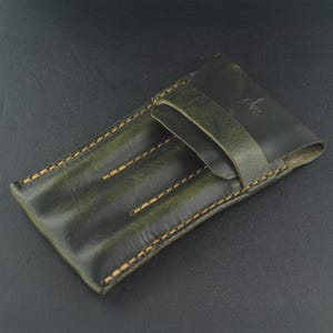 Op de afbeelding: Donkergroene leren etui met drie sleuven, beige stiksels en een klepsluiting. De etui heeft een distressed afwerking en een riemsluiting. Het leer heeft een natuurlijke nerf. Ontworpen voor pennen of potloden.