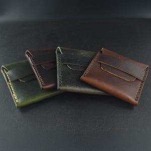 Puede incluir: Cuatro carteras de cuero en tonos verdes, marrones y burdeos se muestran sobre una superficie oscura. Cada cartera tiene un cierre de solapa y costuras visibles. Las carteras varían en color y textura, mostrando las variaciones naturales del cuero.