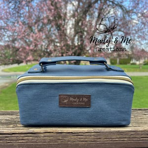 May include: A denim-blue cosmetic bag with a gold zipper and a white accent. It has a handle and a brown leather patch with the text "Mady & Me Creations". The bag is rectangular and sits on a wooden surface.