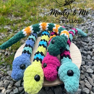 May include: A collection of colorful crocheted snake toys in shades of green, blue, yellow, pink, and teal. Each snake has a black eye and is displayed on a wooden surface. The image includes the text "Mady & Me Creations".