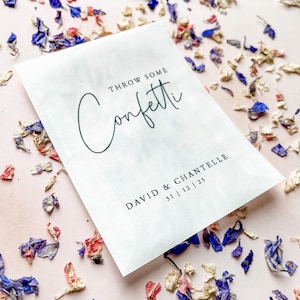 May include: White paper bag with black text that reads "Throw some confetti" and "David & Chantelle 31|12|25". The bag is surrounded by colorful flower petals.