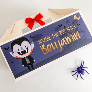 May include: A white cardboard box with a Halloween theme. The box has a cut-out window and a red bow. The box has a blue background with a cartoon vampire and the text "Beware this box bites Ben jamin".