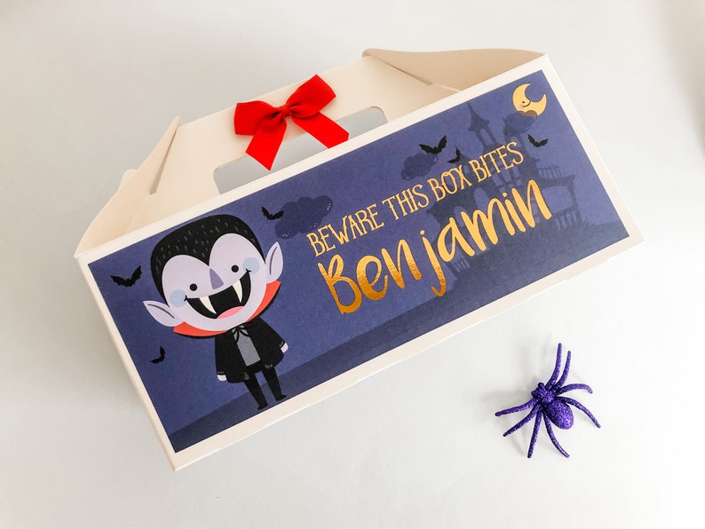 May include: A white cardboard box with a Halloween theme. The box has a cut-out window and a red bow. The box has a blue background with a cartoon vampire and the text "Beware this box bites Ben jamin".