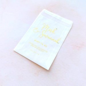 May include: A translucent white paper envelope featuring gold foil lettering that reads Mark & Savannah, MINT TO BE, THANK YOU FOR SHARING OUR SPECIAL DAY, and the date 05.06.25. The envelope has a rectangular shape with a flap at the top.