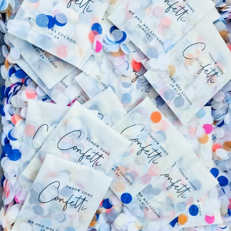 May include: A pile of white paper bags filled with colorful confetti. The bags have the text "Throw Some Confetti" printed on them, along with the names "Mr. and Mrs. Luckoo" and the date "28.07.24".
