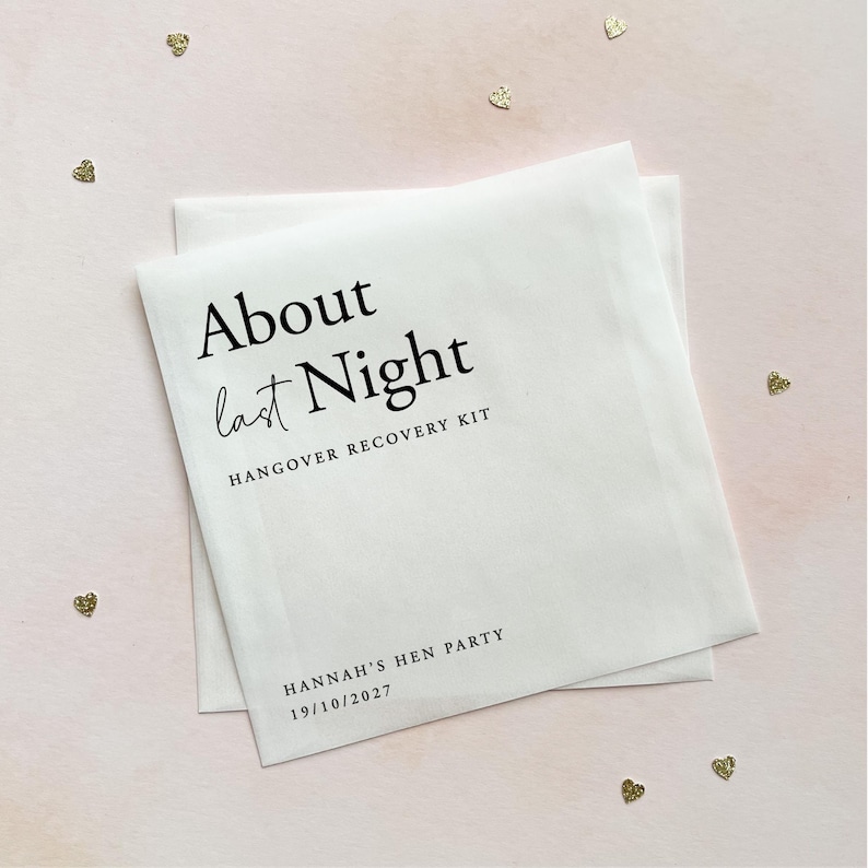 May include: A white square paper envelope featuring black printed text that reads About last Night HANGOVER RECOVERY KIT, followed by HANNAH'S HEN PARTY and the date 19/10/2027. The envelope is surrounded by small gold heart-shaped confetti.