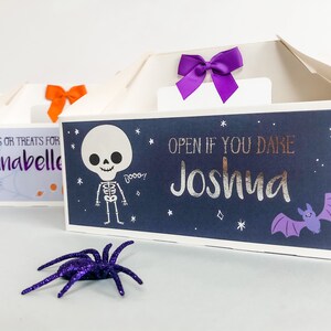 May include: Two Halloween treat boxes with a skeleton and bat design. The boxes are decorated with purple and orange bows. The text on the boxes reads "TRICKS OR TREATS FOR Annabelle" and "OPEN IF YOU DARE Joshua".