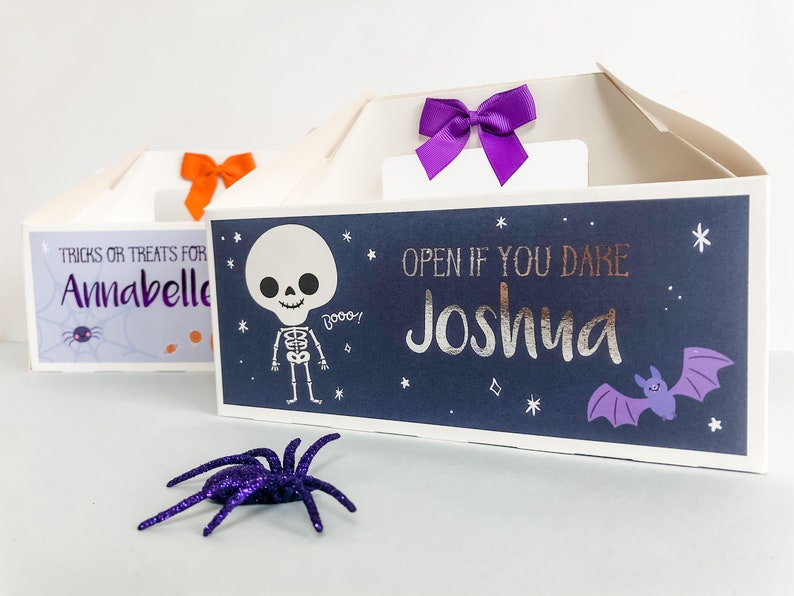 May include: Two Halloween treat boxes with a skeleton and bat design. The boxes are decorated with purple and orange bows. The text on the boxes reads "TRICKS OR TREATS FOR Annabelle" and "OPEN IF YOU DARE Joshua".