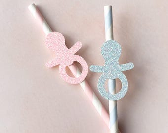 May include: Two paper straws with baby-themed decorations. One straw has a pink glitter pacifier, and the other has a blue glitter pacifier. The straws have pink and white, and blue and white stripes, respectively. The background is a solid light pink.