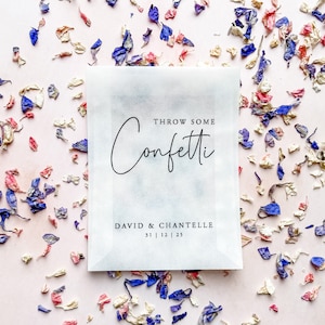 May include: White paper bag with the text "Throw Some Confetti" and "David & Chantelle 31 | 12 | 25" printed on it. The bag is surrounded by dried flower petals in shades of pink, white, and blue.