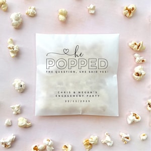 May include: A square, semi-transparent white paper bag filled with popcorn, featuring black text that reads: he POPPED THE QUESTION, SHE SAID YES! CHRIS & MEGAN'S ENGAGEMENT PARTY 22/11/2025. The bag is surrounded by scattered pieces of popcorn.