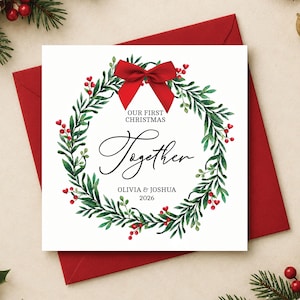 May include: A square white greeting card featuring a watercolor green foliage wreath with red berries and a large red bow at the top. The text reads OUR FIRST CHRISTMAS Together OLIVIA & JOSHUA 2026 in black lettering centered within the wreath.