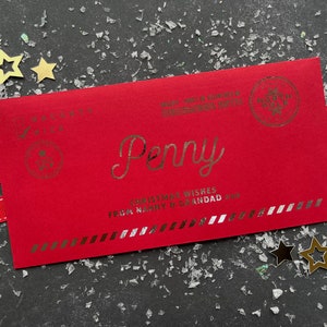 May include: A rectangular red envelope featuring gold foil text that reads Penny, Christmas Wishes From Nanny & Grandad xxx, and Urgent - Must Be Delivered By December 25th. It includes a North Pole stamp, a Tiny Elf seal, and a Naughty or Nice checklist.