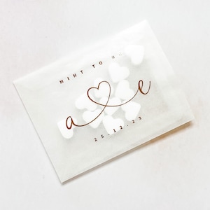 May include: White paper bag with the words "Mint to be" and a gold heart design. The bag is filled with white heart-shaped confetti.