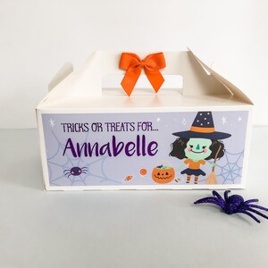 May include: A white cardboard box with a handle and an orange bow on top. The box has a purple and white design with a cartoon witch, a spider, and the text "TRICKS OR TREATS FOR... Annabelle".