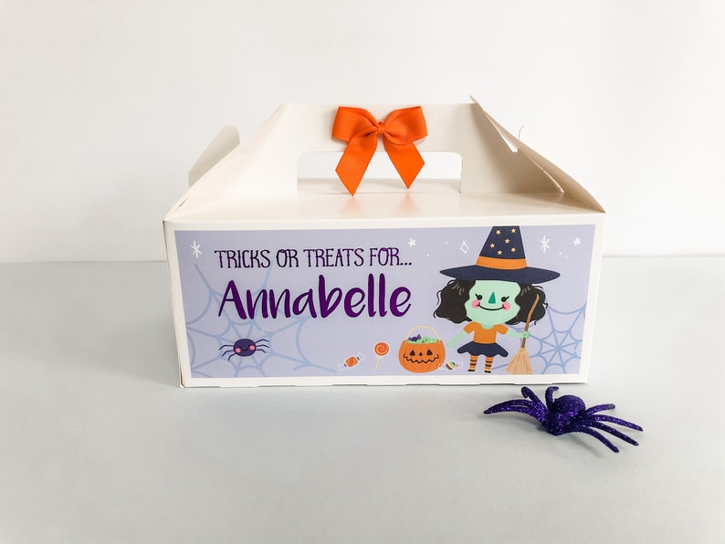 May include: A white cardboard box with a handle and an orange bow on top. The box has a purple and white design with a cartoon witch, a spider, and the text "TRICKS OR TREATS FOR... Annabelle".