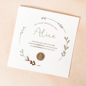May include: A square white card featuring a circular botanical illustration in grey and brown. The text reads: ON YOUR WEDDING DAY, Alice, SOMETHING OLD, SOMETHING NEW, SOMETHING BORROWED, SOMETHING BLUE & A LUCKY SIXPENCE FOR YOUR SHOE. A metal coin is attached.