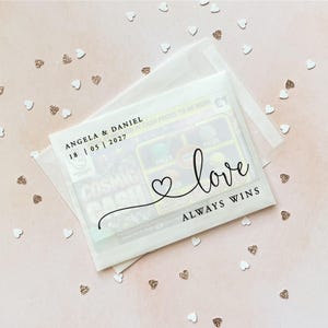 May include: A clear, translucent envelope with the words "love always wins" in black script. The envelope contains a lottery ticket and is decorated with small heart-shaped confetti. The names "Angela & Daniel" and the date "18 | 05 | 2027" are printed on the envelope.