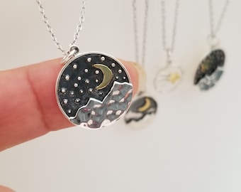 Puede incluir: Un collar de plata con un colgante redondo que presenta una luna creciente y estrellas sobre un fondo negro. El colgante también tiene un diseño de cordillera de plata texturizado.