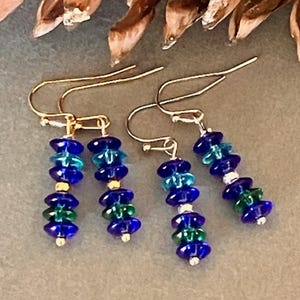 May include: Two pairs of dangle earrings featuring gold-tone ear wires. Each earring consists of a vertical stack of four faceted glass beads in alternating shades of deep blue, teal, and green, separated by small metallic gold-tone spacers.