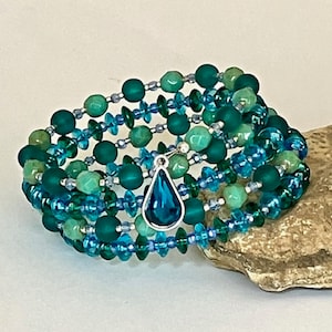 May include: A multi-strand wrap bracelet featuring a variety of beads in shades of teal, turquoise, and emerald green. The design includes matte round beads, faceted crystal beads, and small silver-toned spacers, finished with a teardrop pendant.