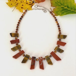 May include: A necklace featuring alternating brown tiger's eye and red jasper rectangular stone segments. The stones are connected by small round brown beads and silver-toned metal spacers, finished with a silver-toned metal chain and clasp.