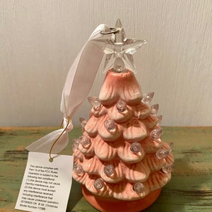 May include: A pink ceramic Christmas tree ornament with a white ribbon and a clear star on top. The tree has clear lights and a label that reads "This device complies with Part 15 of the FCC Rules. Operation is subject to the following two conditions: (1) this device may not cause harmful interference, and (2) this device must accept any interference received, including interference that may cause undesired operation. 20180825 OK Mr. Christmas Model Number: 17399"