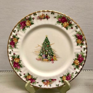 May include: A white porcelain plate with a gold rim, featuring a Christmas tree scene. The plate is decorated with a floral border of red and yellow roses. The plate is displayed on a green stand.