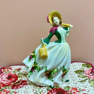 May include: A porcelain figurine of a woman in a green dress and a straw hat, holding a basket. She is standing on a floral patterned fabric.