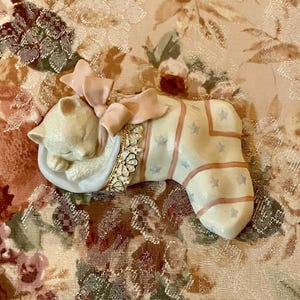 May include: A porcelain figurine of a white kitten sleeping inside a pink and blue stocking with a pink bow and a jeweled trim.