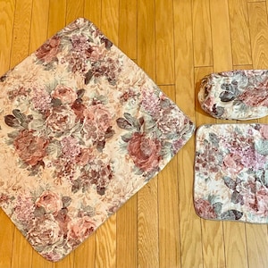 May include: A floral pattern bath set featuring a large square mat, a rectangular mat, and a small oval-shaped mat. The pattern includes pink, red, and green flowers on a cream background. The set is designed for bathroom use.