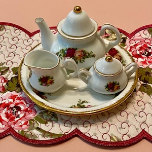 May include: A miniature white porcelain tea set with gold trim, featuring a teapot, creamer, and sugar bowl on a matching tray. Each piece is decorated with a floral rose pattern. The set is displayed on a floral patterned placemat.
