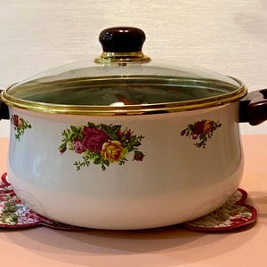 May include: White enamel pot with a glass lid and gold trim. The pot features floral designs in red, yellow, and pink. It has dark brown handles and sits on a floral trivet.