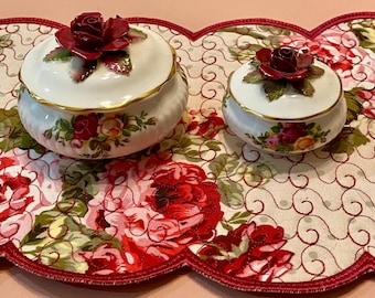 Könnte beinhalten: Zwei weiße Keramik-Schmuckdosen mit goldenem Rand und rosenförmigen Deckeln. Die Dosen sind mit floralen Mustern verziert und liegen auf einem Stoff mit floralem Muster und einem gewellten Rand. Die Rosen auf den Deckeln sind tiefrot.
