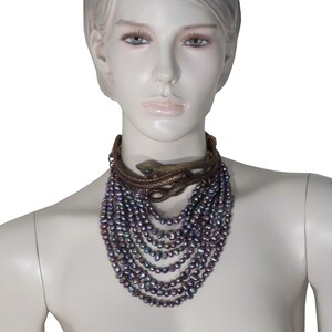 Puede incluir: Un collar de m&uacute;ltiples hilos con perlas de color p&uacute;rpura iridiscente y un colgante de serpiente de oro. El collar est&aacute; en capas y cuelga hasta el pecho.