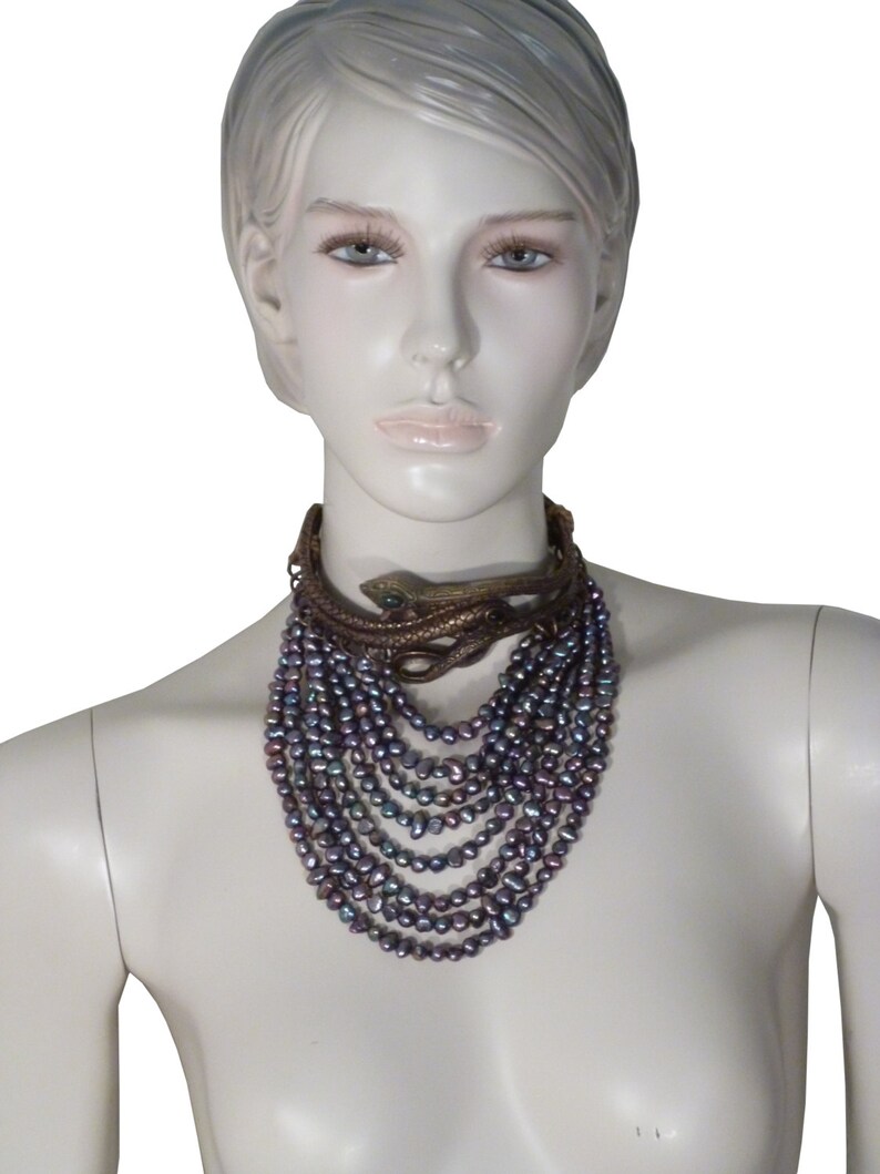 Puede incluir: Un collar de m&uacute;ltiples hilos con perlas de color p&uacute;rpura iridiscente y un colgante de serpiente de oro. El collar est&aacute; en capas y cuelga hasta el pecho.