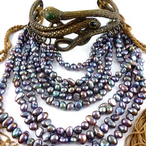 Puede incluir: Un collar de m&uacute;ltiples hilos hecho de perlas negras iridiscentes con un brazalete de serpiente de lat&oacute;n con piedras verdes y negras.