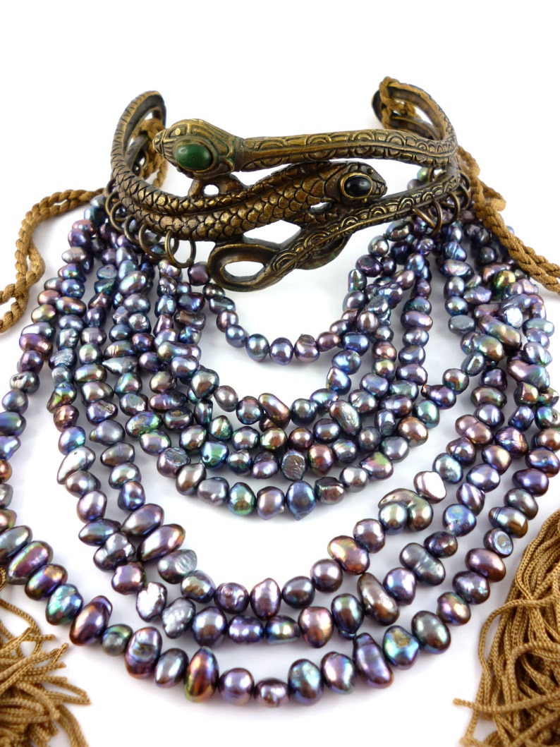 Puede incluir: Un collar de m&uacute;ltiples hilos hecho de perlas negras iridiscentes con un brazalete de serpiente de lat&oacute;n con piedras verdes y negras.