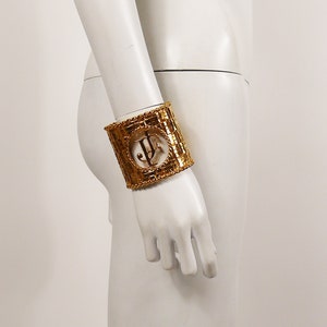 May include: Gold-toned metal bracelet with a woven design and a large, open, circular cutout featuring the letters "JF".