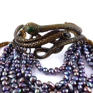Puede incluir: Un brazalete de doble serpiente dorado con piedras verdes y negras, combinado con un collar de m&uacute;ltiples hilos de perlas de color p&uacute;rpura oscuro.