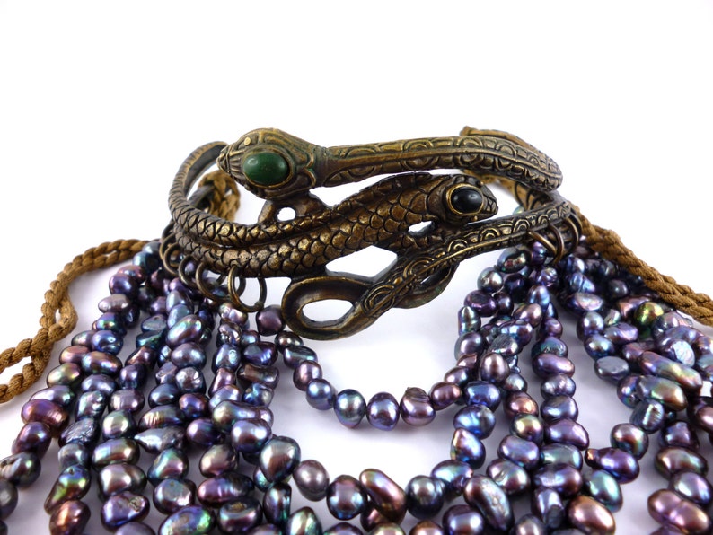 Puede incluir: Un brazalete de doble serpiente dorado con piedras verdes y negras, combinado con un collar de m&uacute;ltiples hilos de perlas de color p&uacute;rpura oscuro.