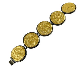 Puede incluir: Un brazalete de oro y negro con cinco eslabones redondos, texturados y dorados. Los eslabones están conectados por metal negro.