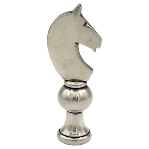 May include: A silver-colored bottle stopper in the shape of a chess knight. The knight's head is detailed with a curved neck and a defined jawline. The stopper has a spherical midsection and a flared base.