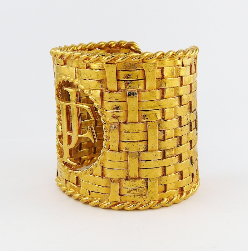 May include: A gold-tone woven cuff bracelet with a large, cutout "F" monogram. The bracelet is made of a thick, textured metal and has a wide, open design.