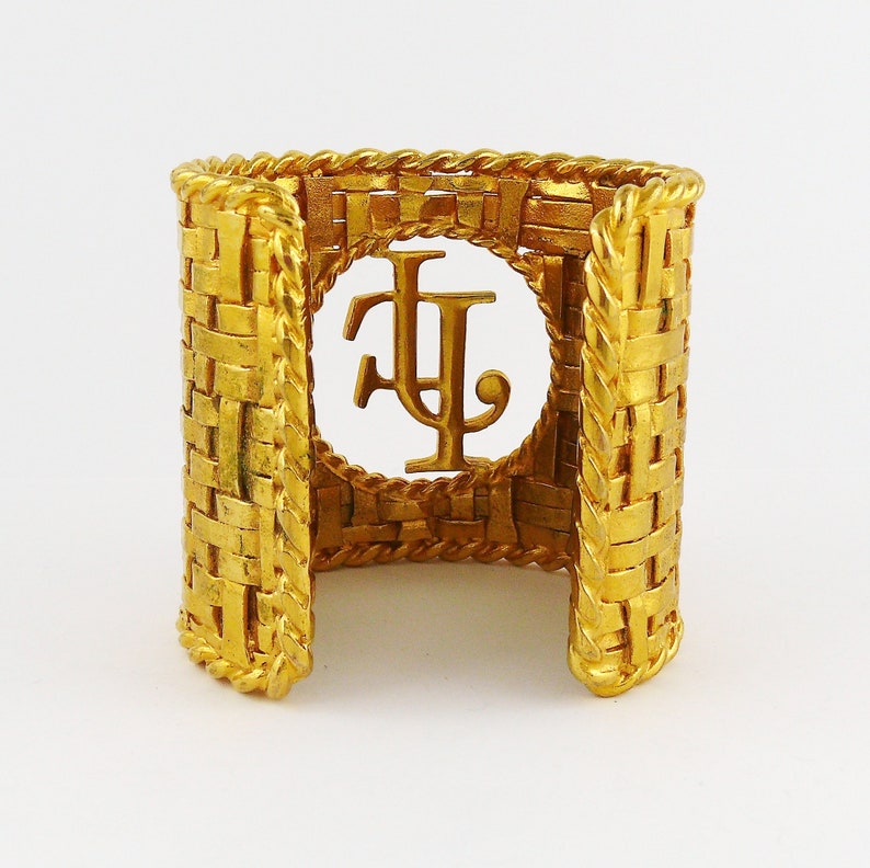 May include: A gold-tone woven cuff bracelet with a large, open, circular design featuring the initials "F.L." in the center.