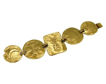 Puede incluir: Un brazalete dorado con un diseño de cadena de eslabones. El brazalete presenta cinco dijes, cada uno con un diseño diferente: una luna creciente, un trébol de cuatro hojas, el logotipo de YSL, un corazón y una estrella.