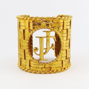 May include: Gold-toned woven cuff bracelet with a large, circular cutout featuring the initials "JF" in a stylized font.