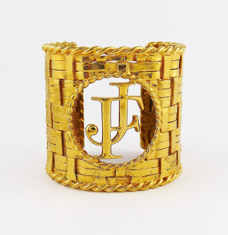 May include: Gold-toned woven cuff bracelet with a large, circular cutout featuring the initials "JF" in a stylized font.